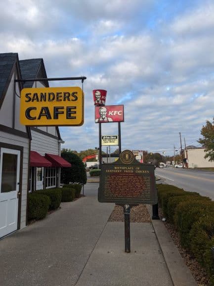 White door with a yellow sign that says 'Sanders Cafe" in black. There is a sign on the sidewalk in front of the building that tells the history of the original KFC building.