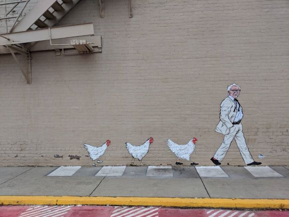 Mural of Colonel Sanders followed by three chickens painted on a cream brick wall. The figures appear to all be walking in a line.