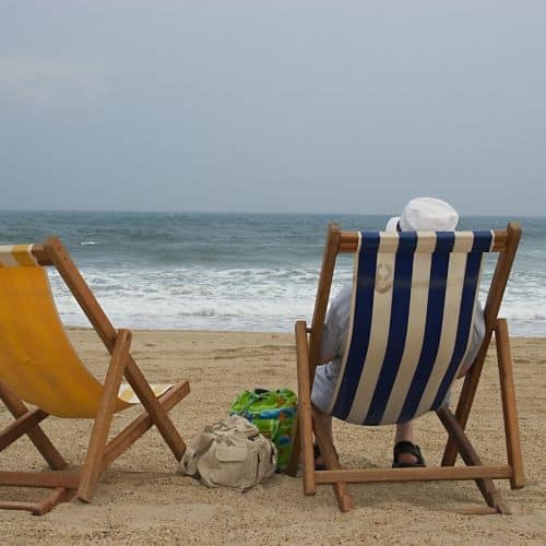 Beach with two beach chairs; one yellow, one blue and white striped. There are two bags between the chair. The striped chair is occupied by a person wearing a white hat.