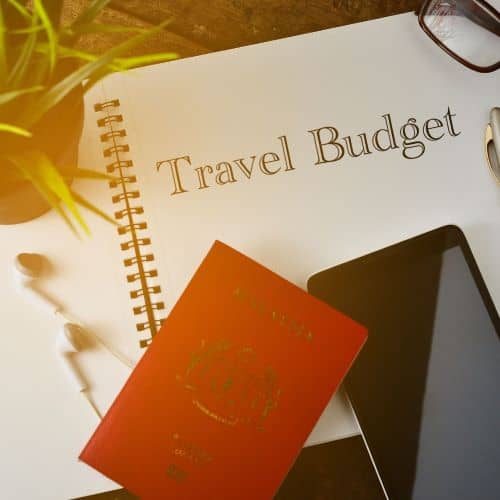 Open notebook with the words "Travel Budget". Towards the bottom is a passport and smart phone.