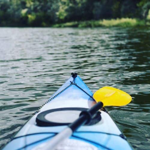 tip of a blue kayak in the water. There is greenery in the distance