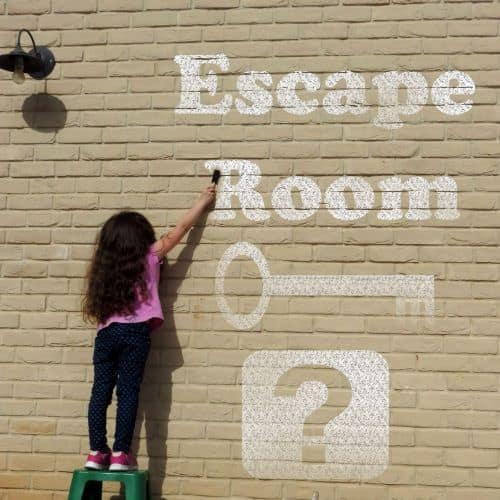 Girl writing "Escape Room" on a wall in chalk. There is also a key and a question mark.