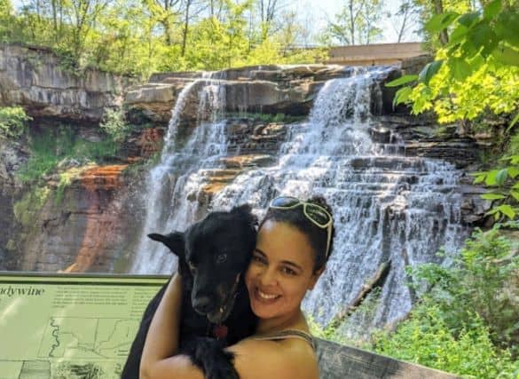 A woman holding her black dog in front of a large waterfall- Brandywine Falls