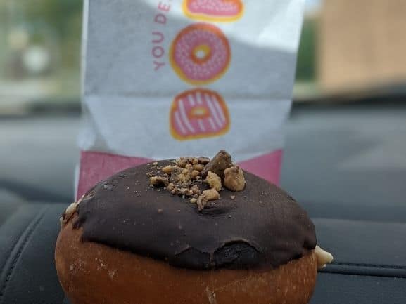 Close up of a Peanut butter and Chocolate donut in front of  a pink and white donut bag. Donut has chocolate frosting and PB crumbles on top.