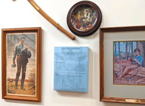 A blue sign that describes the "Railsplitter" Scene in the Lincoln Museum. It is placed in between two drawings of Abe Lincoln as a child and as a young man. Above the sign is an axe and another drawing of Abe Lincoln as a young man.