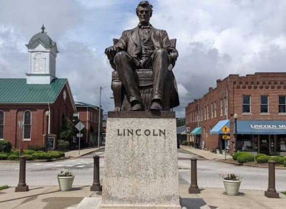 Statue of Abe Lincoln sitting in a chair on top of a stone pedestal that says "Lincoln" You can see the Lincoln Museum in the background. It is in the middle of downtown Hodgenville.
