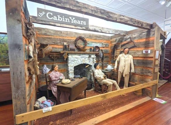 A scene from the Lincoln Museum. There is a sign that says "the Cabin Years" at the top. The scene is of a young Abe Lincoln sitting in a rocking chair in between his parents. They are in a log cabin.