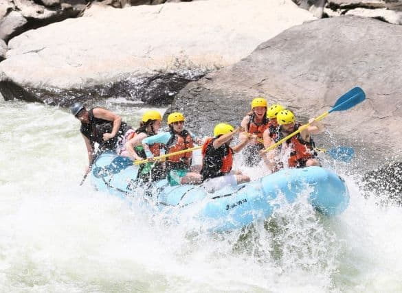 Group of 6 friends white water rafting in Lansing West Virginia. They are mid rapid with water spray all around them and front of boat is midair
