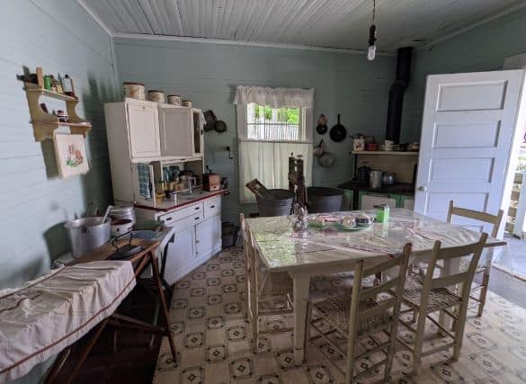 The kitchen of a miner's family home. There is a table, a stove, an ironing table and two wash bins for laundry.