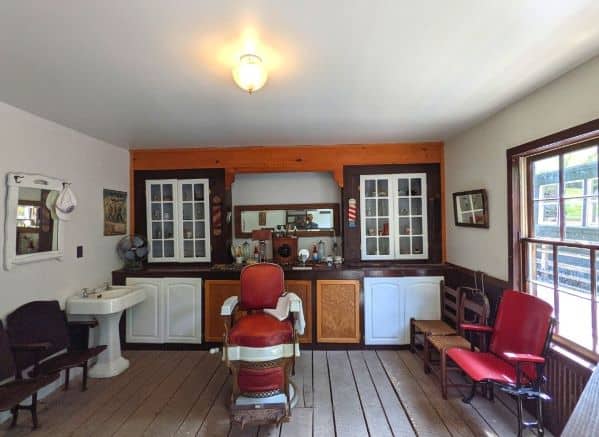 A room set up like an old barber shop/medical office. There is a chair in the middle with benches along side the wall. There is also a free standing sink on the back left side of the room and a counter with various equipment along the back wall