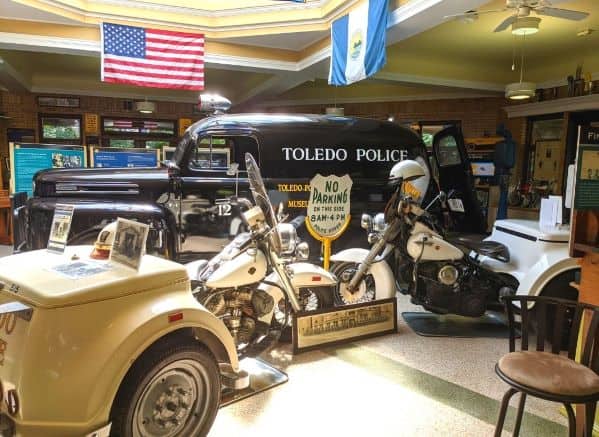 2 old-fashioned police motorcycles displayed in front of an old-fashioned Toledo Police car inside the Toledo Police Museum