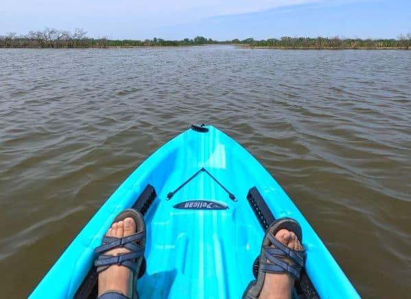 2 feet wearing sandals at the front of a blue kayak. The kayak is in the middle of a body of water