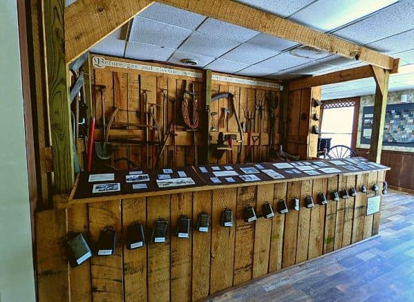 A display made of wood to look like a counter. There are farming tools against the back wall and on the front of the counter is a row of all different types of bells