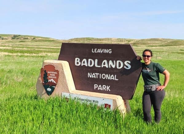 Woman wearing grey leggings and a green shirt standing next to a "Leaving Badlands National Park" sign in a prairie field