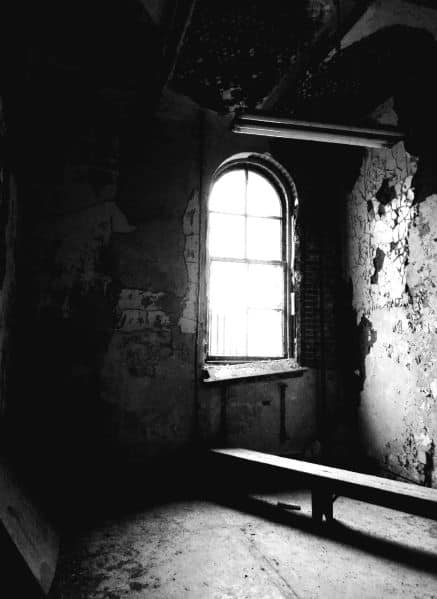 Black and white photo of a dilapidated corner of the Reformatory. Paint is peeling off of the wall and debris is on the floor next to an abandoned wooden bench