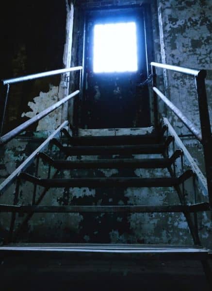 View looking up a metal staircase to a closed chained door. Light is coming in through the barred window in the door. Paint is peeling off of the walls