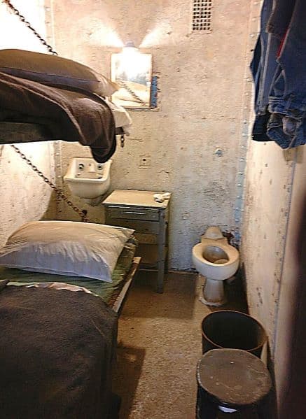 photo of a prisoner's cell in the Reformatory. There is a bunk bed, sink, toilet, and side table with a jean jacket hanging on a wall