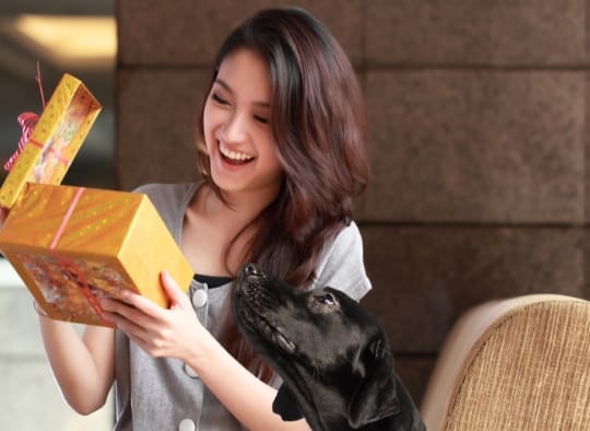 Woman opening a gift with her dog looking up at it 
