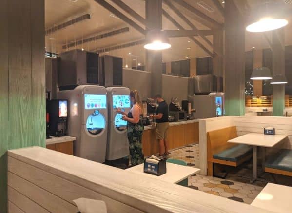 Multiple self-serve soda machines, hot chocolate dispenser, microwave, and several condiment areas all along a counter.