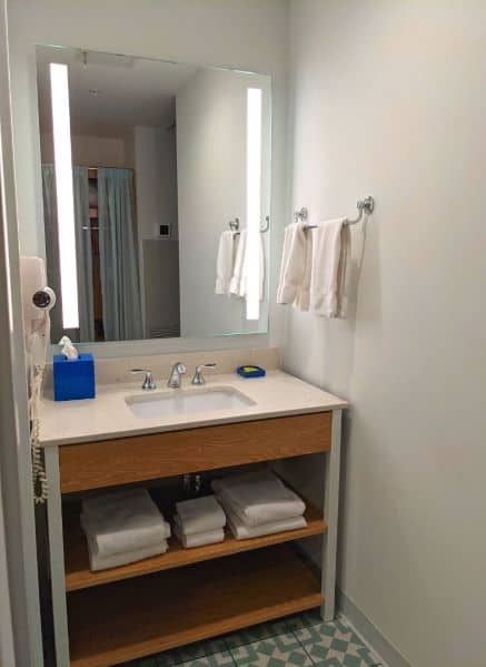 Sink area outside bathroom. There are two shelves below sink with extra towels. Facial tissues and a blow dryer are next to sink and mirror
