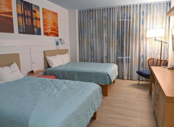 Standard room in Dockside Inn & Suites. There are 2 queen sized beds with blue coverlets on left side. 3 tropical pictures above beds. Right side has a chair and long shelf/desk area. Back wall is all windows.