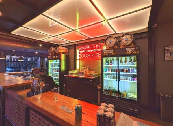 Bar at the Check-in Counter for DogHouse Hotel. Warm ambient lighting fills the room contrasted with a red neon sign.  Two glass front refrigerators shine brightly while displaying an assortment of colorful drinks in many sizes and shapes. Two beer keys adorn the top of each fridge.
