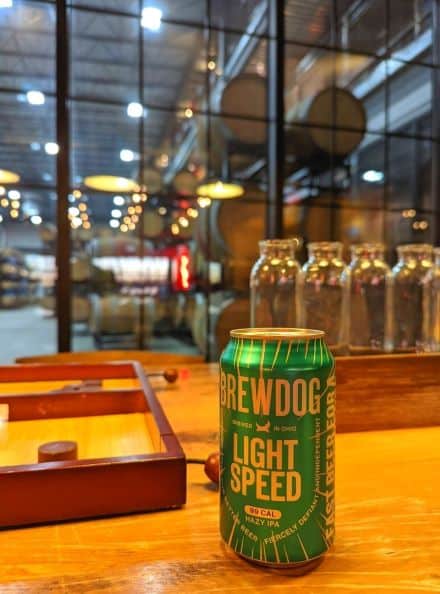 Green beer can next to a wooden table game in the lobby of DogHouse Hotel. Behind both items is a enormous glass window ridged with steel. Behind window , hundreds of varying sizes of wooden fermenting barrels stacked three floors high can be seen.