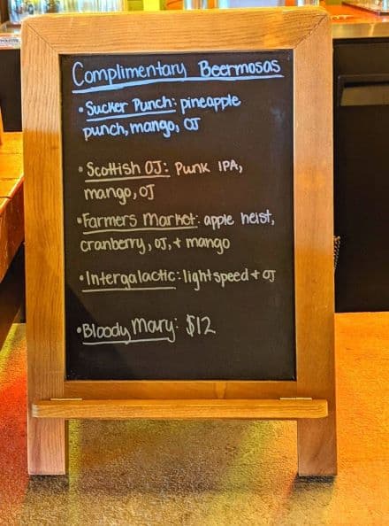 The menu for the complimentary beermosas. Menu is on a rectangular black chalkboard with a wooden frame. Menu items are written in chalk paint . Both the colors blue and white are used in menu text.