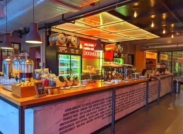 Continental breakfast spread in DogHouse Hotel. Beverage containers, fruit, yogurt toppings, cereal, pastries, and more. Warm ambient lighting fills the room  contrasted with a red neon sign behind the extended L shaped bar kiosk. 