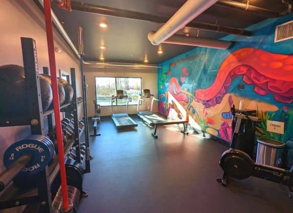 DogHouse Fitness center. There are a wall of weights, treadmills, and a stationary bike. Right wall contains a colorful mural of blues, pinks, reds, greens portraying a large octopus tentacle.