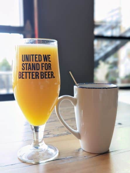 A  close up shot of a glass of Beermosa that says "United we stand for better beer" next to a mug of coffee on a wooden table.