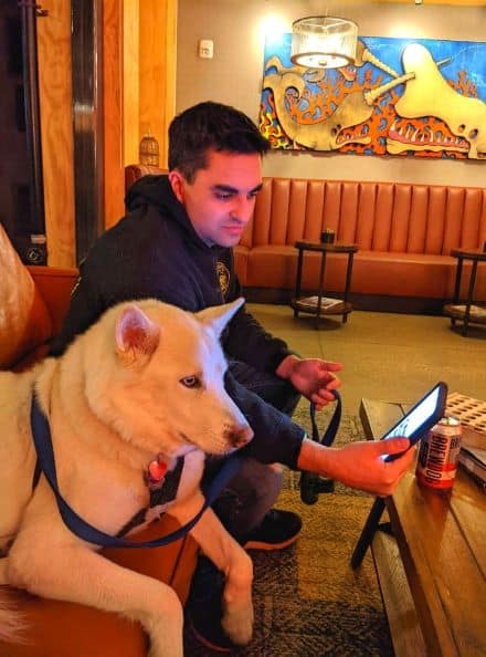 Man sitting next to a white husky on a couch and table with beer in the hotel lobby of DogHouse Hotel for Happy Hour. Man is showing dog images on a google pixel 6. Warm ambient lighting fills the room. A  large art piece can be seen behind the man and dog. The art piece consist of narwhals fighting in front of fire. Art piece is made of wood. Blues, reds and pinks color the piece as well as the natural grain of the wood.