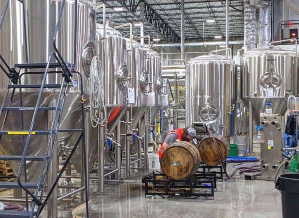 Man wearing an orange vest kneeling by wooden barrels in the middle of BrewDog Brewery. Man is surrounded by industrial sized brewing supplies and machines.