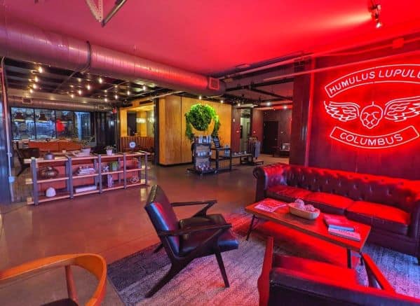 One of the seating area in the DogHouse Hotel Lobby, there are couches and chairs. A large red neon sign is on wall that has an image of a hop with wings and is surrounded by text that reads HUMULUS LUPULUS, COLUMBUS. The light from the sign casts a reddish hue amongst the chairs and table.