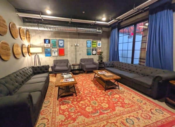 TV room at DogHouse Hotel. Several couches and two chairs surround two rectangular wooden coffee tables. All these items are placed on a large red rug. Various framed art adorn the walls. A large steel framed window  can be seen on right side of room which has a view into fermenting room. 