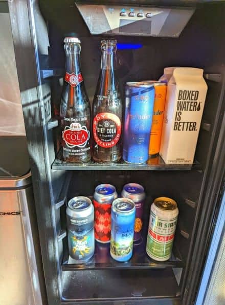 Selection of water, soda, and beer in a mini fridge inside hotel room at DogHouse. A range of drinks fill fridge from glass bottles, tall colorful cans to cardboard boxed water . 