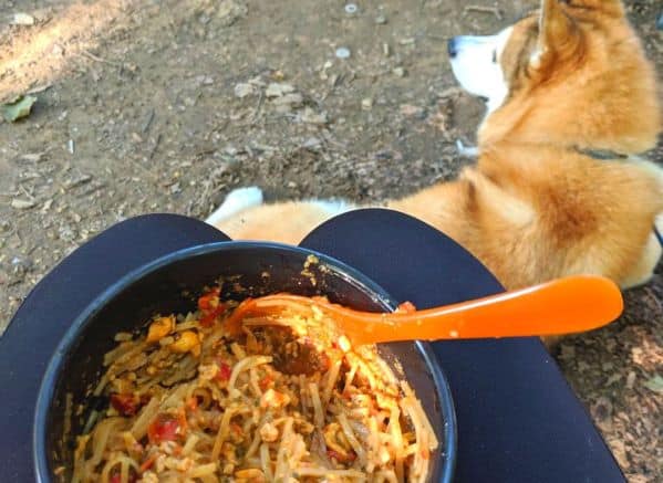 bowl of camping food on a person's lap with a dog laying down in the background.