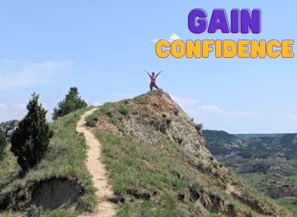 Gain Confidence written in sky next to woman standing on top of mountain ledgs