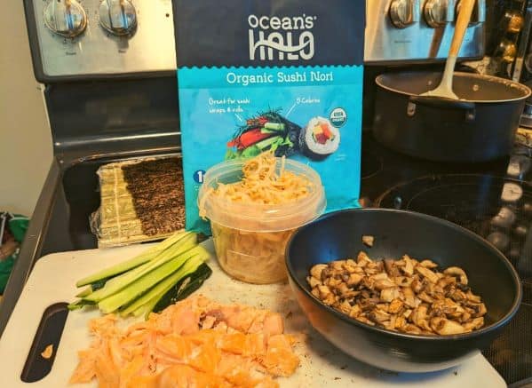 Cutting board with smoked salmon, bean sprouts, cucumbers, mushrooms on it with a bag of seaweed wraps in back