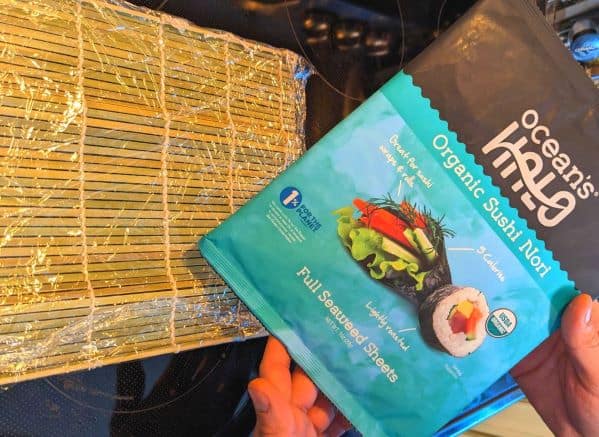 Seaweed wraps being held in front of a sushi rolling mat wrapped in plastic wrap