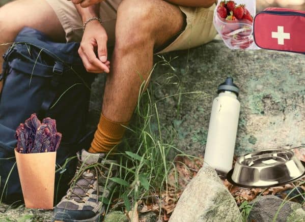 person sitting on a rock next to a hiking bag, bag of jerky, first aid kit, bag of strawberries, and a dog water bowl