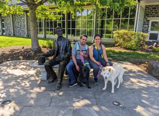 Couple with their 3 dogs of various sizes sitting on bench next to an Abe Lincoln statue in front of Gettysburg Visitor Center