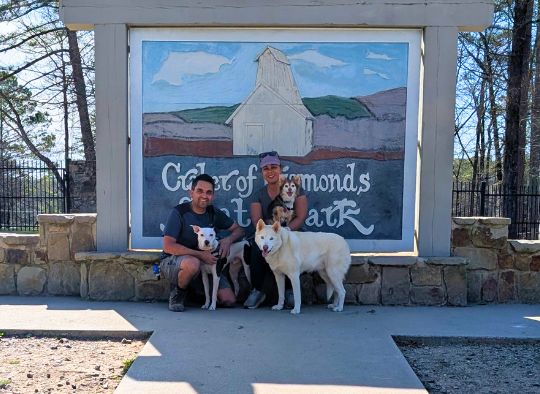 3 dogs and their 2 owners in front of the Crater of Diamonds State Park sign