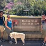 3 people, each with a dog standing next to Hot Springs National park sign