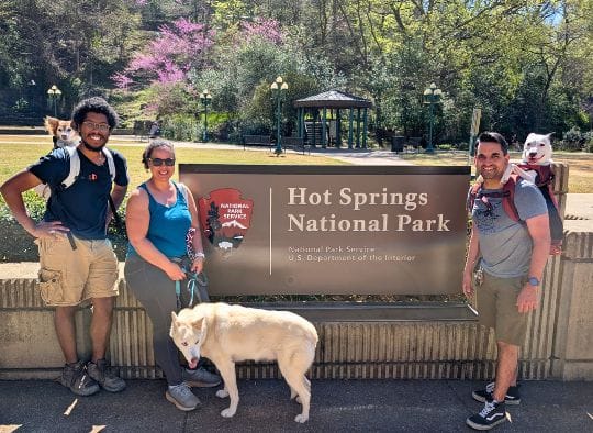 3 people, each with a dog next to the Hot Springs National Park sign