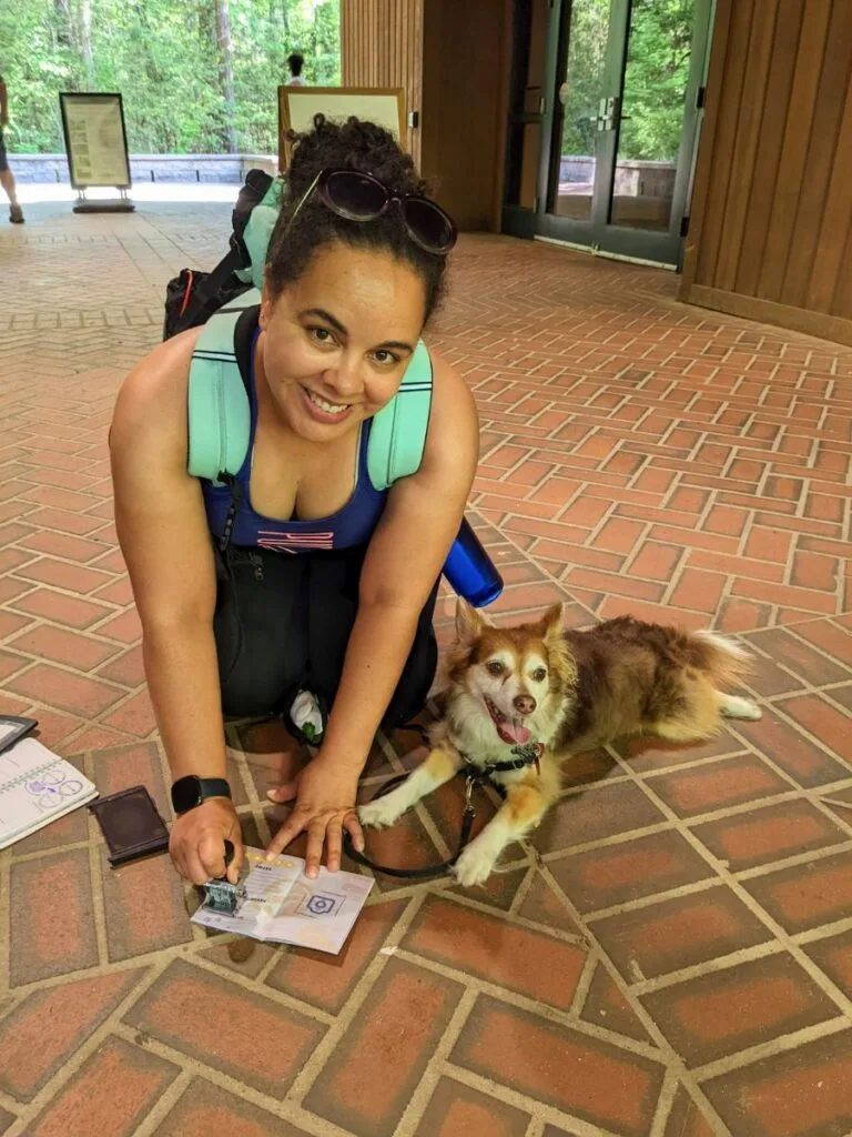 Woman kneeling next to small dog stamping Pup Memory Passport in Congaree National Park