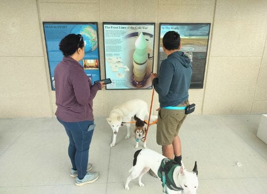 2 people with 3 dogs looking at an outdoor sign at dog friendly Minuteman Missile Silo NHS